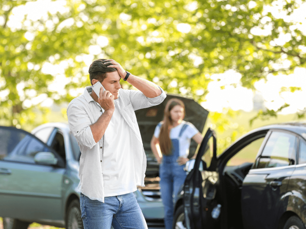 Man holding head and talking on phone near two cars after accident; woman stands by open car door and popped hood in background, illustrating the first steps after a car accident.